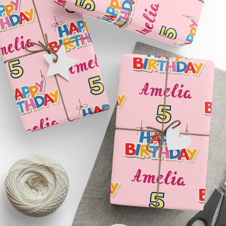 May include: Pink birthday wrapping paper with colourful text that reads "Happy Birthday Amelia 5". The gifts are tied with twine and adorned with white paper cutouts. A spool of twine and scissors are also visible.