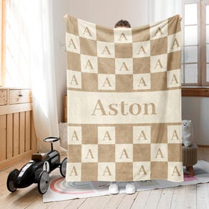 May include: A beige and brown checkered blanket with the name "Aston" printed in the center. The blanket has the letter "A" in each square. The blanket is being held up by a person.