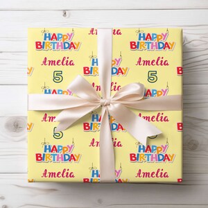 May include: A wrapped present with yellow birthday wrapping paper. The paper is patterned with the words "Happy Birthday," the name "Amelia," and the number "5." A cream ribbon is tied in a bow around the gift.
