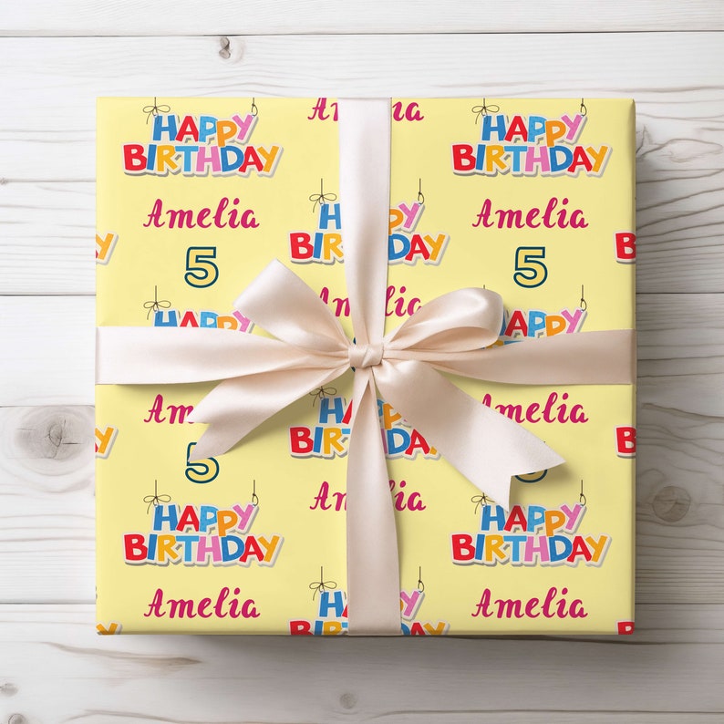 May include: A wrapped present with yellow birthday wrapping paper. The paper is patterned with the words "Happy Birthday," the name "Amelia," and the number "5." A cream ribbon is tied in a bow around the gift.