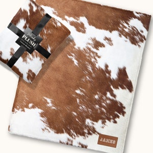 May include: A brown and white cowhide-patterned blanket with a matching folded blanket tied with a black ribbon. The folded blanket has a tag that reads "Minky Plush Custom Blanket". A leather tag with the name "JAMES" is sewn on the blanket.