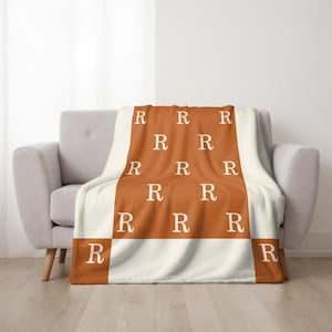 Puede incluir: Una manta suave y mullida con un patrón repetido de la letra "R" en blanco sobre un fondo naranja quemado. La manta está doblada y drapeada sobre un sillón gris claro.