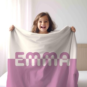 May include: A personalized blanket with the name "EMMA" in large, white letters against a pink and cream background. The blanket is held up by a smiling child.