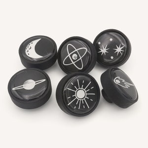 May include: Six black round knobs with different space-themed designs in white. The designs include a crescent moon, an atom, a star, a planet, a sun, and a comet.