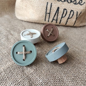 Könnte beinhalten: Vier Holz-Knöpfe mit einem kreuzförmigen Design aus Schnur. Die Knöpfe sind in den Farben Braun, Weiß, Grün und Blau lackiert.