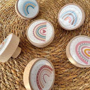 Puede incluir: Seis pomos de madera con diseños de arcoíris. Cada pomo presenta un arcoíris diferente con varios colores y patrones, incluyendo rosa, azul, verde, amarillo y naranja. Los pomos están colocados sobre un fondo tejido.