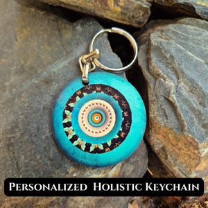 May include: A circular keychain featuring a vibrant turquoise outer ring, a black band with small butterfly silhouettes, and a central mandala-style pattern in shades of beige, orange, and blue. The metal ring is attached to the top of the disc.