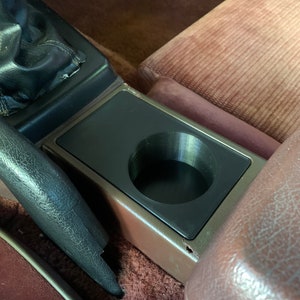 Pode incluir: Um porta-copos de plástico preto está montado no console central de um carro. O porta-copos é rodeado por tecido marrom e um apoio de braço de couro preto.