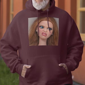 Puede incluir: Una sudadera con capucha granate con un bolsillo delantero y una capucha con cordón. La sudadera presenta una imagen cuadrada de una muñeca con cabello rojo y una expresión de disgusto. La persona que lleva la sudadera sonríe.
