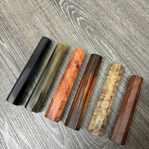 May include: Six rectangular wooden blocks arranged in a row, each featuring a distinct finish. The collection includes a solid black piece, a translucent horn-like piece, a vibrant orange-red burl, a dark wood with vertical grain, a light tan burl, and a deep reddish-brown wood.
