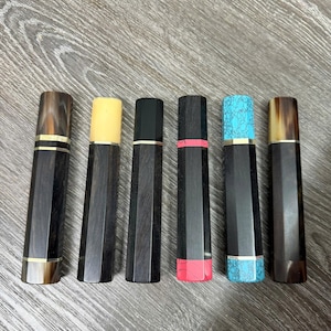 May include: Six knife handles with dark wood bodies and various accent colours. The handles feature gold, yellow, black, pink, and turquoise accents. The handles are arranged in a row on a wooden surface.