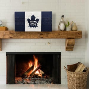 Peut inclure: Une enseigne en bois avec le logo des Maple Leafs de Toronto en bleu et blanc. L'enseigne est suspendue au-dessus d'une cheminée avec un entourage en brique. La cheminée est allumée avec un feu.