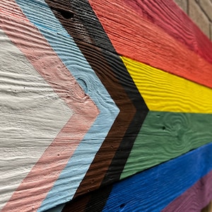 May include: A wooden sign painted with the colours of the Progress Pride flag. The flag is made up of seven stripes: white, pink, light blue, brown, black, light green, blue, and purple. The stripes are arranged in a chevron pattern.