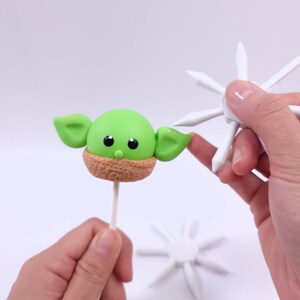 May include: A green, round cake pop resembling a character from a popular science fiction series. The cake pop has large ears, black eyes, and a brown textured collar. A hand holds a white, star-shaped decoration, and another hand holds the cake pop.