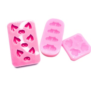 May include: Three pink silicone molds for making resin crafts. One mold has six heart shapes with a small circle in the center of each heart. Another mold has four cloud shapes. The third mold has four flower shapes.