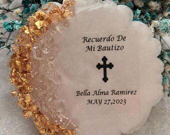 May include: A circular white resin coaster with a scalloped edge, featuring a crescent of gold leaf flakes on the left side. The centre displays black text reading Recuerdo De Mi Bautizo, a black cross icon, the name Bella Alma Ramirez, and MAY 27, 2023.