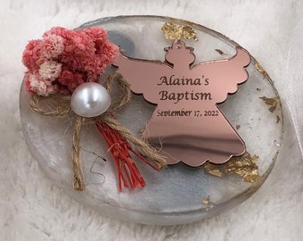 May include: A circular white resin coaster with gold leaf flakes, featuring a rose gold mirror angel ornament engraved with "Alaina's Baptism" and "September 17, 2022". A cluster of pink dried flowers, a white pearl, and twine are attached to the side.