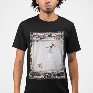 May include: A black t-shirt with a graphic print of a boxing match. The image shows two boxers in a ring, with a crowd of people surrounding the ring. The text "ALI" is visible in the image.