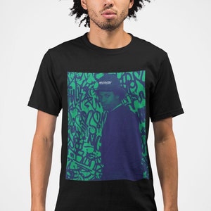 Puede incluir: Una camiseta negra con un estampado gráfico de una persona con un sombrero de pie frente a una pared de graffiti verde y azul. El texto "WESTSIDE" está en el sombrero.
