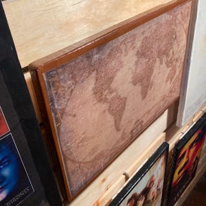 May include: A framed world map with a vintage style design. The map is printed on a light brown paper with a distressed look. The frame is a dark brown wood with a light brown border.
