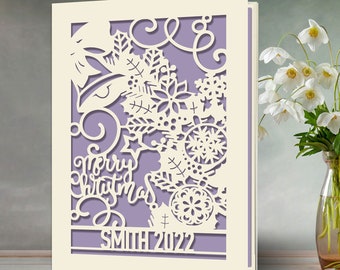May include: A cream-colored Christmas card with a laser-cut design. The design includes snowflakes, ornaments, and the words "Merry Christmas" and "SMITH 2022" on a lavender background. The card is standing upright.