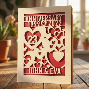 May include: A cream-colored anniversary card with a red interior. The card features laser-cut hearts and the words "ANNIVERSARY 1999 2022" and "23" along with the names "JOHN & EVA". The card is displayed on a wooden surface.