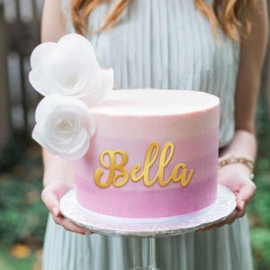 May include: A pink ombre cake with the name "Bella" written in gold script on top. The cake is decorated with two white paper flowers.