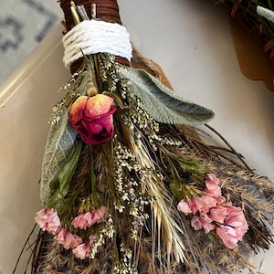 May include: A decorative bundle of dried botanicals featuring a central pink rosebud, light pink statice flowers, sage leaves, and wheat stalks. The stems are tightly bound with brown twine and wrapped with a thick white cord at the top handle.