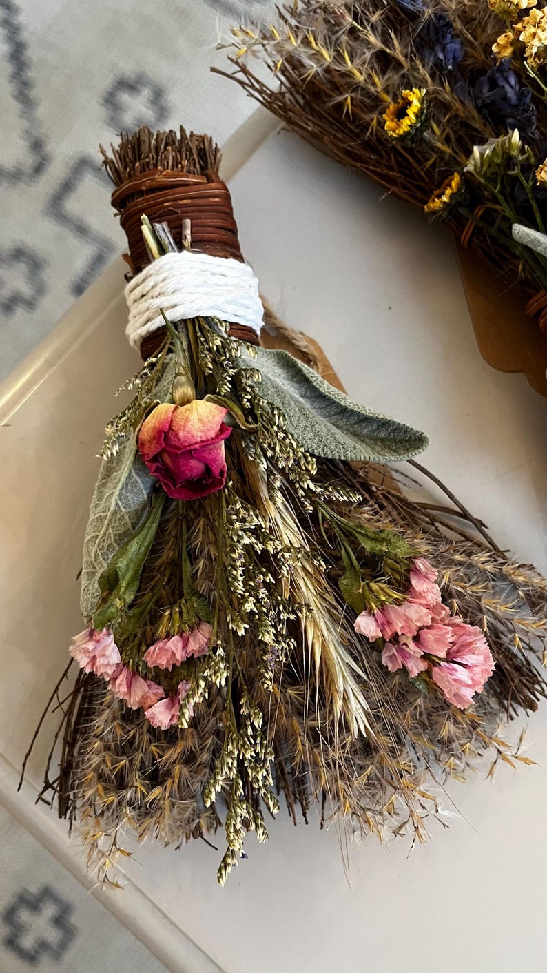 May include: A decorative bundle of dried botanicals featuring a central pink rosebud, light pink statice flowers, sage leaves, and wheat stalks. The stems are tightly bound with brown twine and wrapped with a thick white cord at the top handle.
