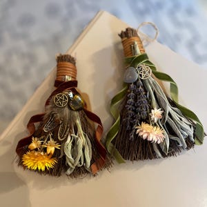 May include: Two decorative brooms with natural bristles, adorned with dried flowers, feathers, and ribbons. One features a yellow flower and brown ribbon, the other lavender and green ribbon. Both have a pentacle charm.