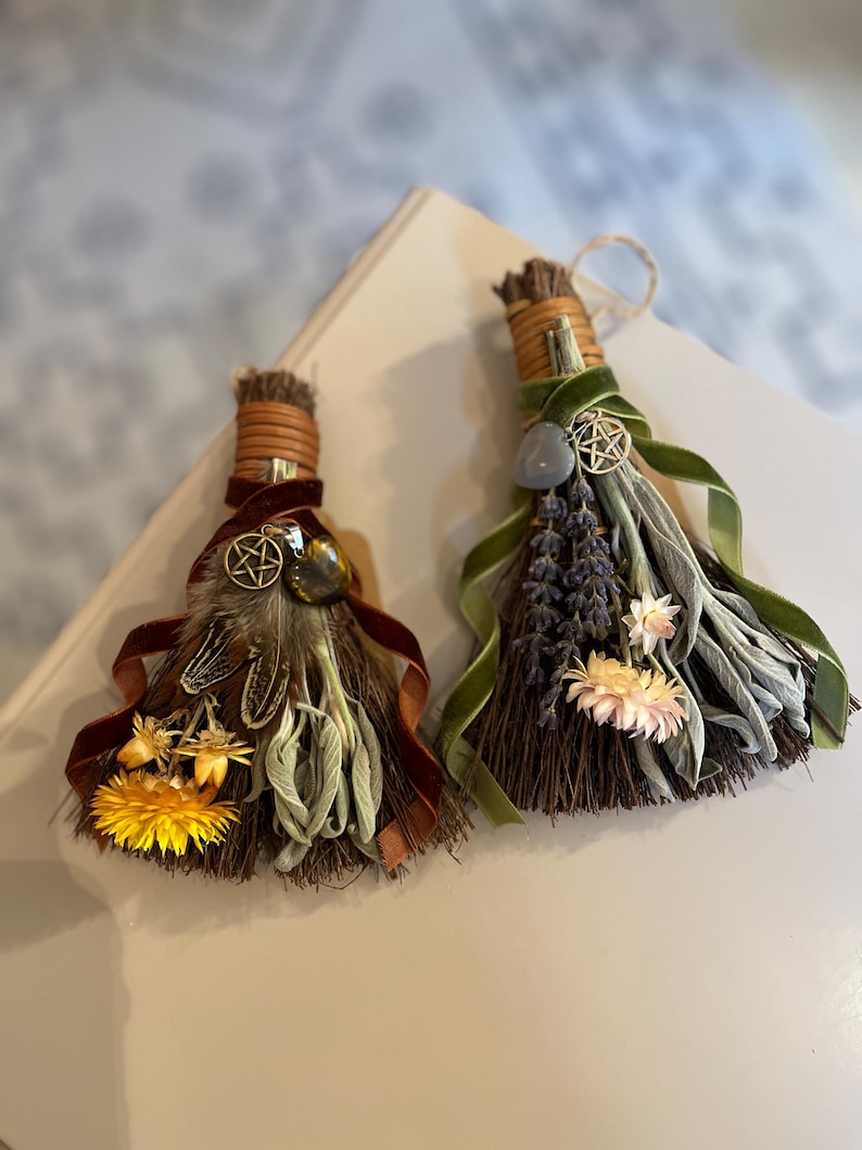 May include: Two decorative brooms with natural bristles, adorned with dried flowers, feathers, and ribbons. One features a yellow flower and brown ribbon, the other lavender and green ribbon. Both have a pentacle charm.