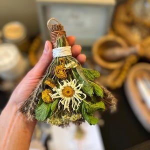May include: A small decorative broom made of dried herbs and flowers, featuring a central white daisy, yellow marigolds, and green mint leaves. The handle is wrapped with brown twine and secured with a white cord, creating a bundle of dried botanicals.