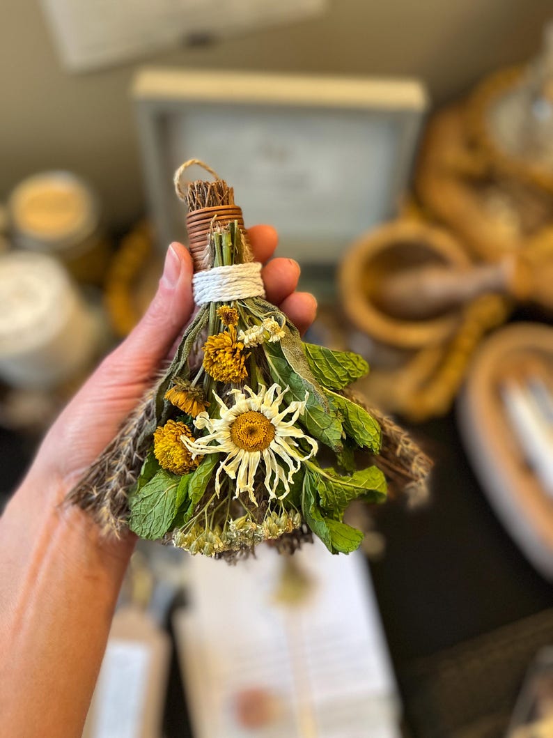 May include: A small decorative broom made of dried herbs and flowers, featuring a central white daisy, yellow marigolds, and green mint leaves. The handle is wrapped with brown twine and secured with a white cord, creating a bundle of dried botanicals.