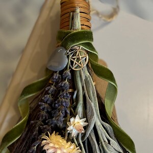 May include: A decorative broom featuring dried lavender, sage, and flowers, wrapped with a green velvet ribbon. A silver pentacle charm and a gray stone are attached. The broom is made of natural materials.