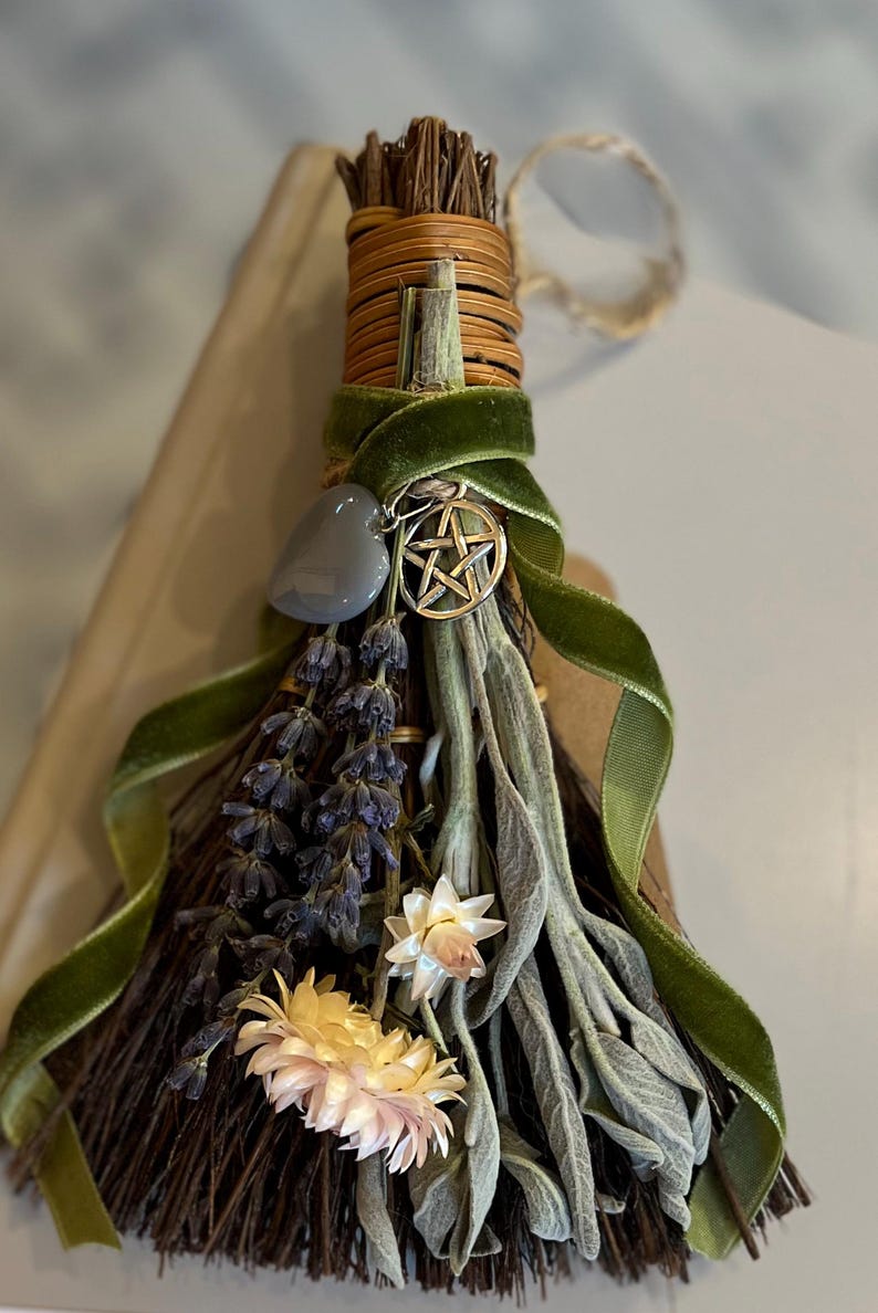 May include: A decorative broom featuring dried lavender, sage, and flowers, wrapped with a green velvet ribbon. A silver pentacle charm and a gray stone are attached. The broom is made of natural materials.