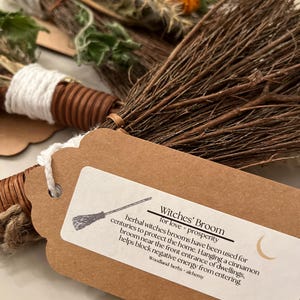May include: A small decorative broom made of thin brown twigs, wrapped with white yarn and tied with a brown paper tag. The tag features a crescent moon icon and text reading: Witches' Broom for love + prosperity, herbal witches brooms have been used for centuries to protect the home. Hanging a cinnamon broom near the front entrance of dwellings, helps block negative energy from entering. Woodland herbs + alchemy.