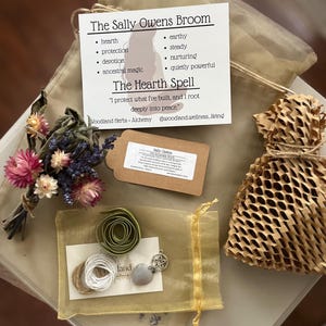 May include: A gift set featuring a card titled The Sally Owens Broom with The Hearth Spell text, a dried flower bouquet, a brown paper tag, a small honeycomb-patterned paper bag, and a gold organza pouch containing green ribbon, white twine, and a pentacle charm.