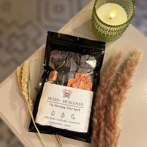 May include: A black resealable pouch labelled Hexed + Hungover The Morning After Spell with a skull and heart icon. Inside are a grey scrunchie, a small orange bottle-shaped item, and stones. A green glass candle and dried grass stalks.