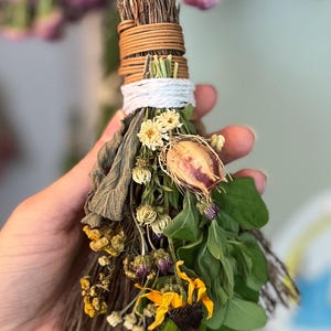 May include: A small broom made of bundled brown twigs, tightly wrapped with tan and white cord at the handle. The broom is decorated with an assortment of dried flowers, including yellow blossoms, green leaves, and a single dried seed pod.