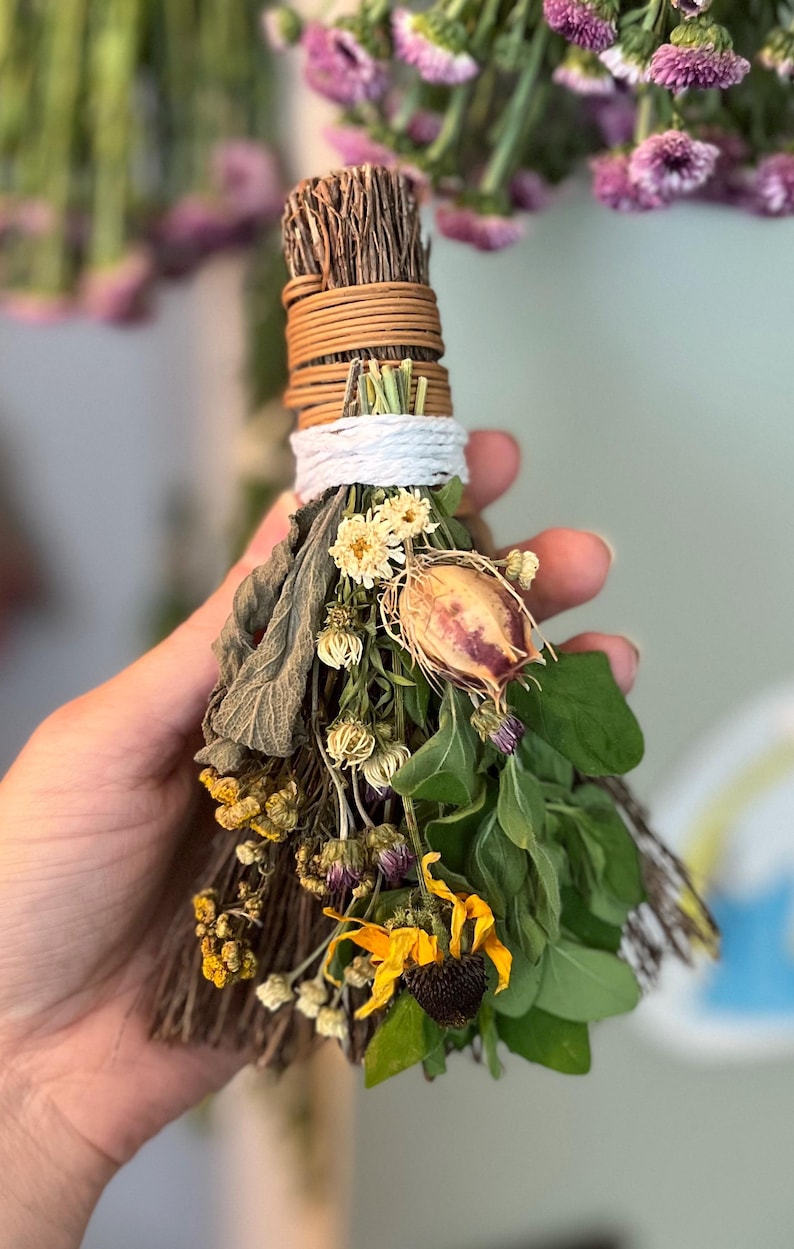 May include: A small broom made of bundled brown twigs, tightly wrapped with tan and white cord at the handle. The broom is decorated with an assortment of dried flowers, including yellow blossoms, green leaves, and a single dried seed pod.