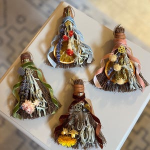 May include: Four decorative miniature brooms made of brown twigs, each adorned with dried flowers, herbs, citrus slices, velvet ribbons, and small metal pentacle charms. The arrangements feature varied color palettes including blue, pink, green, and dark red accents.