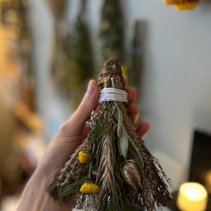 May include: A small, triangular broom made of dried brown fibres, bound at the top with white string and a brown leather cord. The front is decorated with dried green leaves, yellow flowers, a stalk of wheat, and a small, dried seed pod.