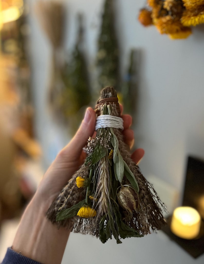 May include: A small, triangular broom made of dried brown fibres, bound at the top with white string and a brown leather cord. The front is decorated with dried green leaves, yellow flowers, a stalk of wheat, and a small, dried seed pod.