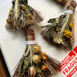 May include: Three small decorative brooms crafted from bundles of dried herbs, grasses, and flowers. Each broom is tied with white string and brown wire, featuring yellow marigold blossoms, seed pods, and various green foliage textures.