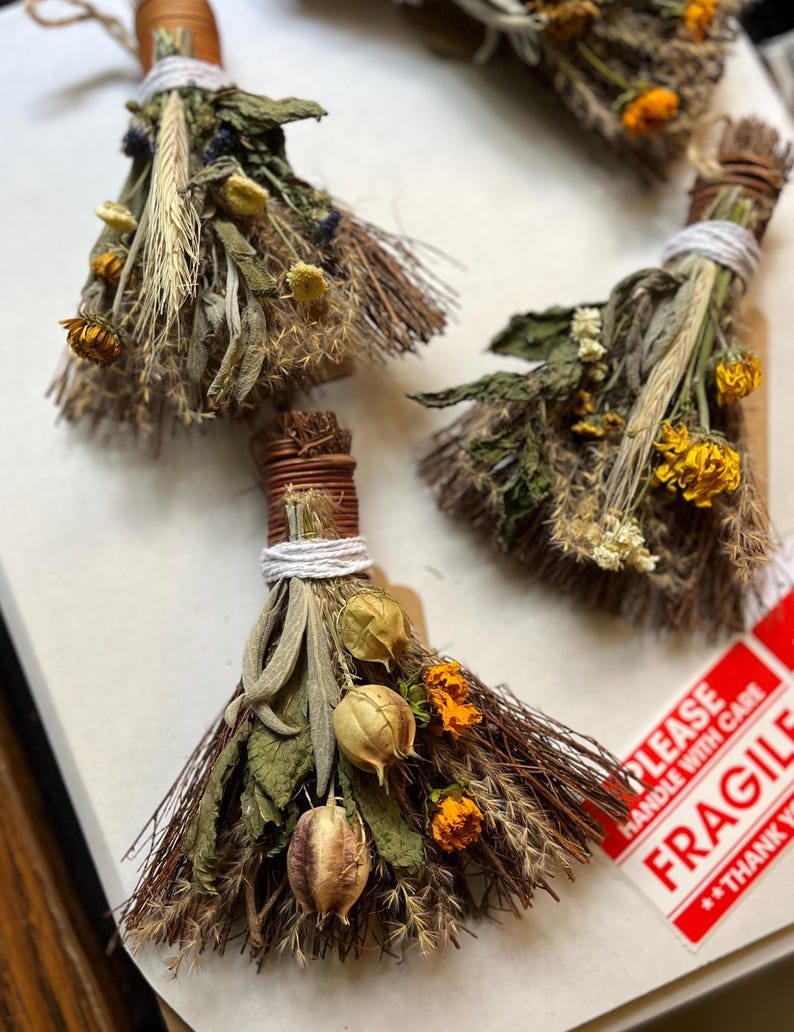 May include: Three small decorative brooms crafted from bundles of dried herbs, grasses, and flowers. Each broom is tied with white string and brown wire, featuring yellow marigold blossoms, seed pods, and various green foliage textures.