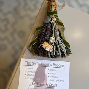 May include: A handcrafted Sally Owens Broom, featuring dried herbs, flowers, and a green velvet ribbon. A gray stone and a gold-colored pentacle charm are attached. The accompanying card reads "The Sally Owens Broom" and "The Hearth Spell."