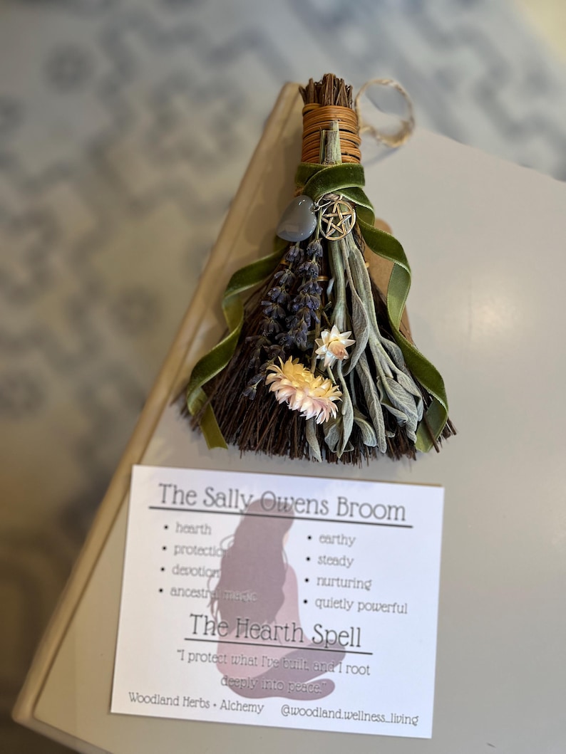 May include: A handcrafted Sally Owens Broom, featuring dried herbs, flowers, and a green velvet ribbon. A gray stone and a gold-colored pentacle charm are attached. The accompanying card reads "The Sally Owens Broom" and "The Hearth Spell."