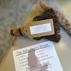 May include: A small, handcrafted broom with a tag that reads "Sally Owens." The broom is wrapped with green velvet ribbon and gold twine. A card with the text "The Sally Owens Broom" and "The Hearth Spell" is also present.