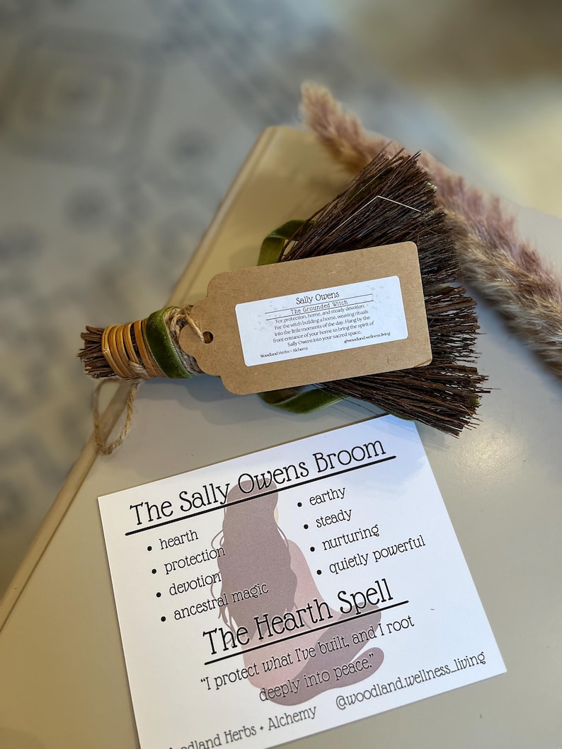 May include: A small, handcrafted broom with a tag that reads "Sally Owens." The broom is wrapped with green velvet ribbon and gold twine. A card with the text "The Sally Owens Broom" and "The Hearth Spell" is also present.