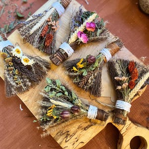May include: Five small decorative brooms made of dark brown twigs, each tied with white or brown string and adorned with various dried flowers, including pink roses, yellow daisies, orange wheat stalks, and green foliage, arranged on a wooden board.